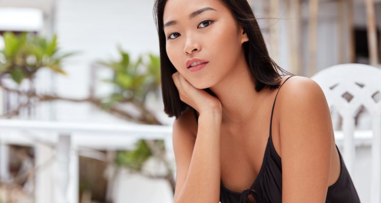 Horizontal shot of serious attractive Asian young female model with dark hair, make up and healthy skin, sits against terrace cafe interior, waits for friend who is late for meeting, feels bored